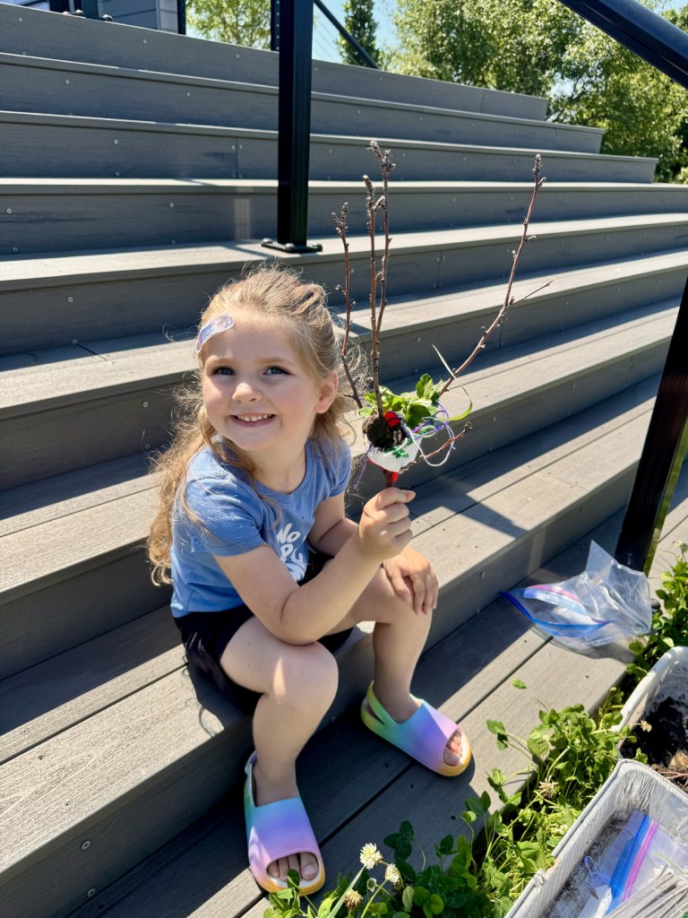 A young girl sitting on steps, smiling while holding a decorated stick with colorful items, surrounded by greenery.
