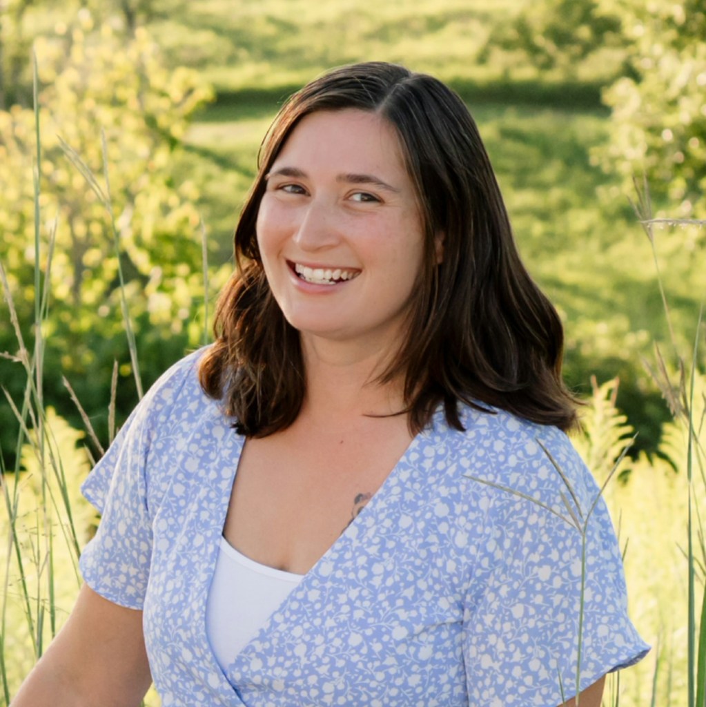 Smiling woman with long brown hair wearing a light blue floral dress, standing among tall grass outdoors.