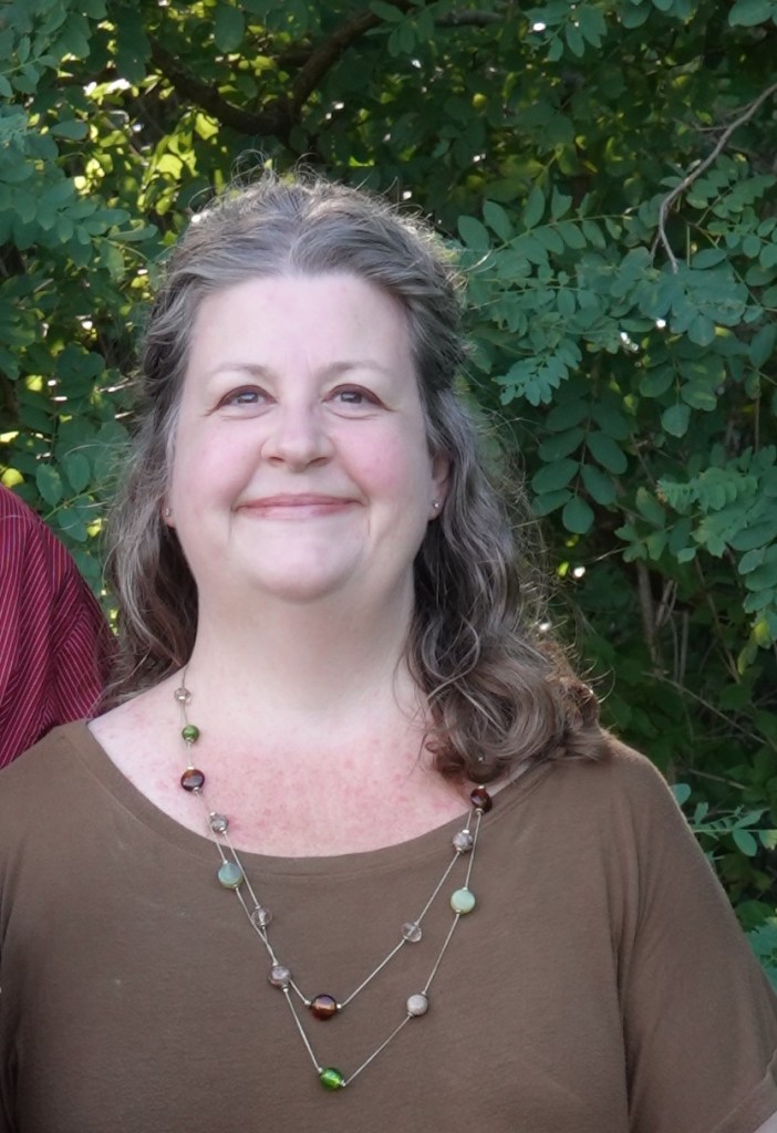 Portrait of a woman with curly, shoulder-length hair, wearing a brown top and a beaded necklace, smiling against a backdrop of green foliage.